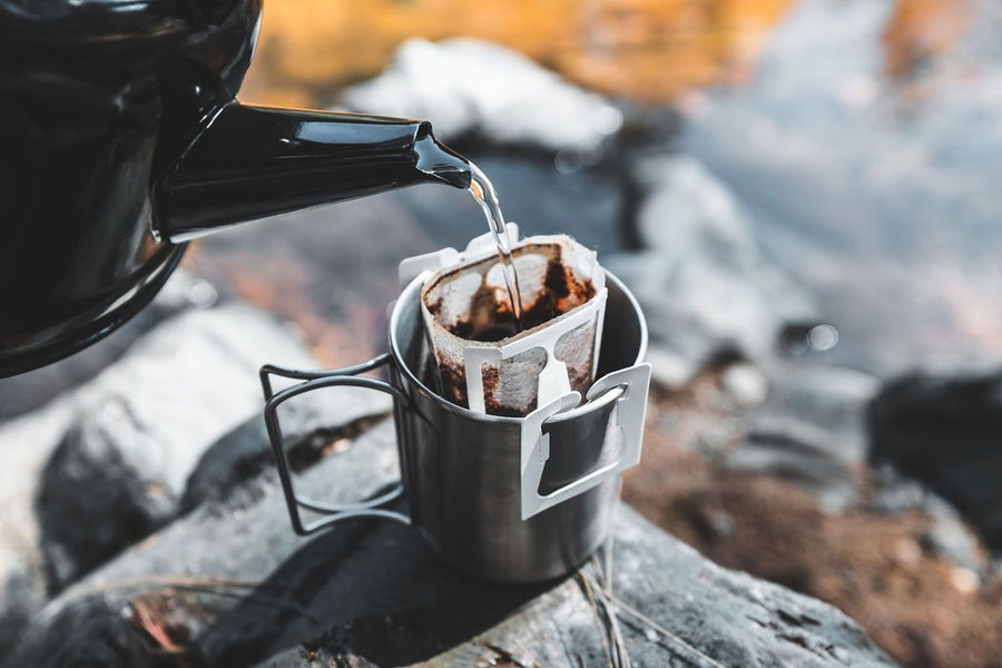 Kettle pouring hot water into a drip coffee bag on a camp cup beside a stream