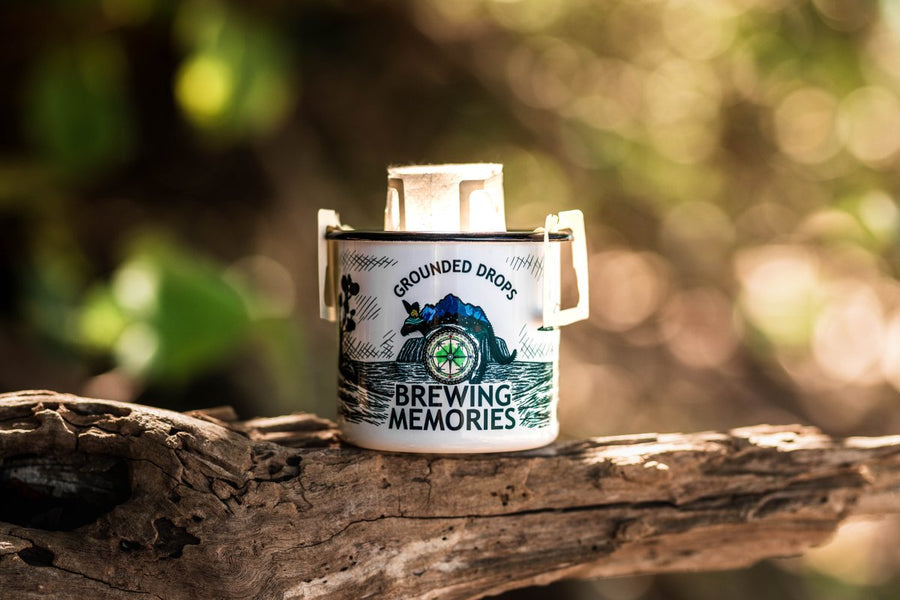 Grounded Drops Brewing Memories enamel mug with a drip coffee bag on a log