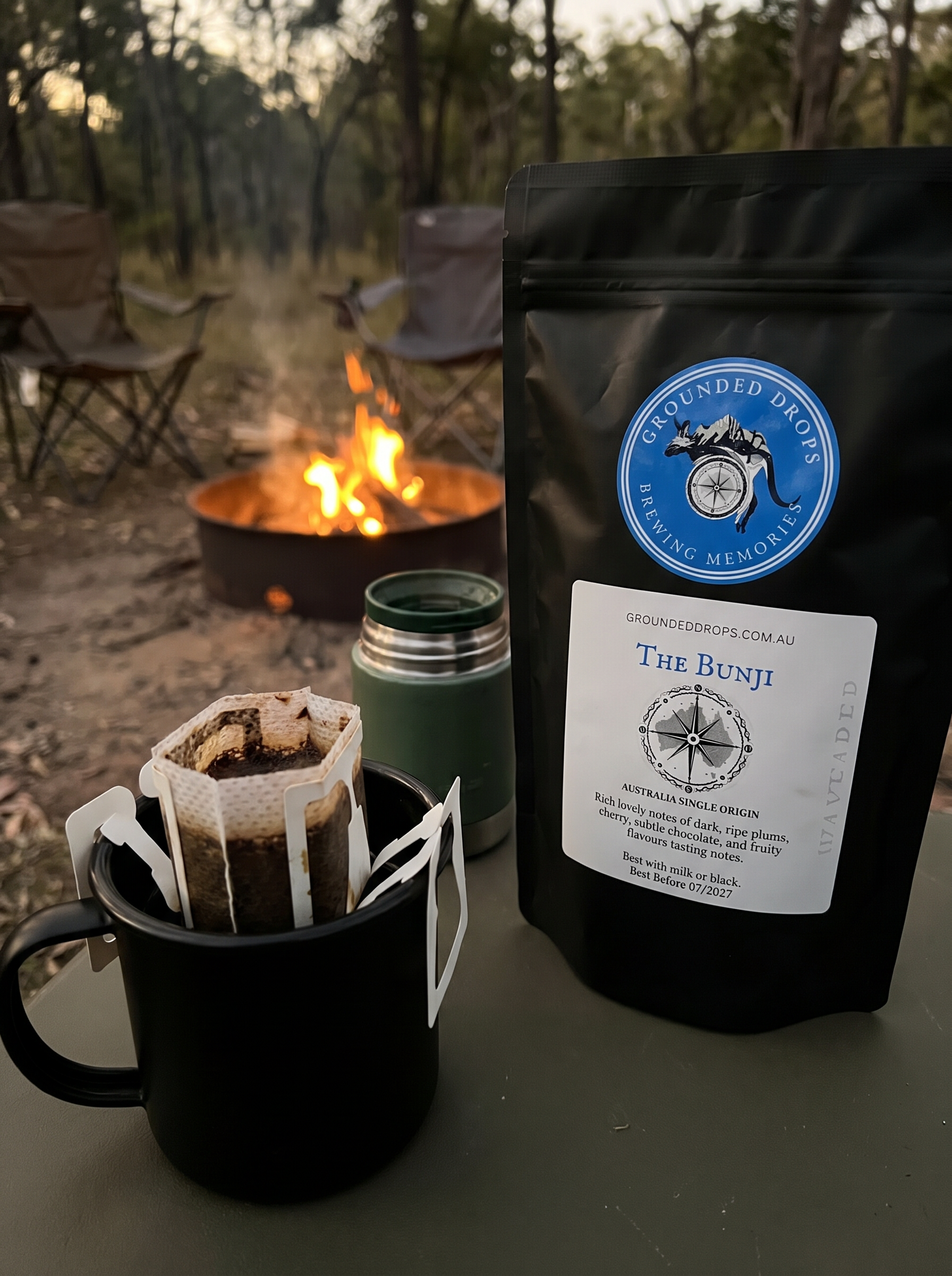 Grounded Drops drip coffee bag brewing in a camp mug beside a campfire at dusk
