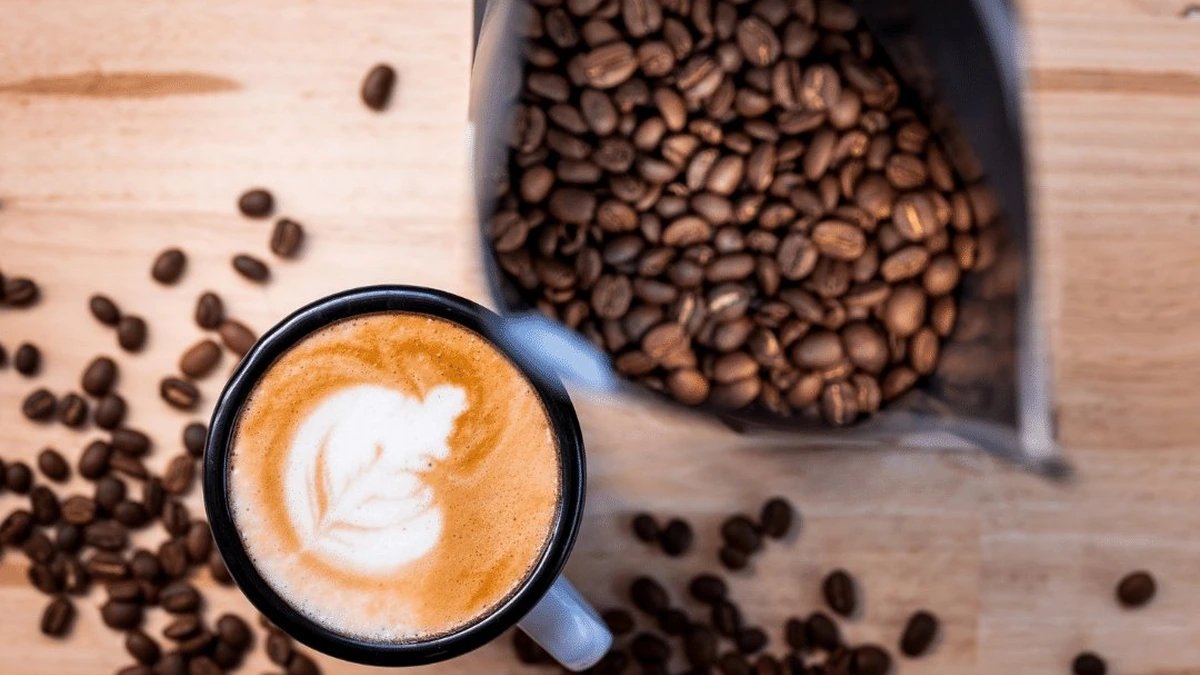 An enamel cup of latte beside an open bag of coffee beans on a timber table.
