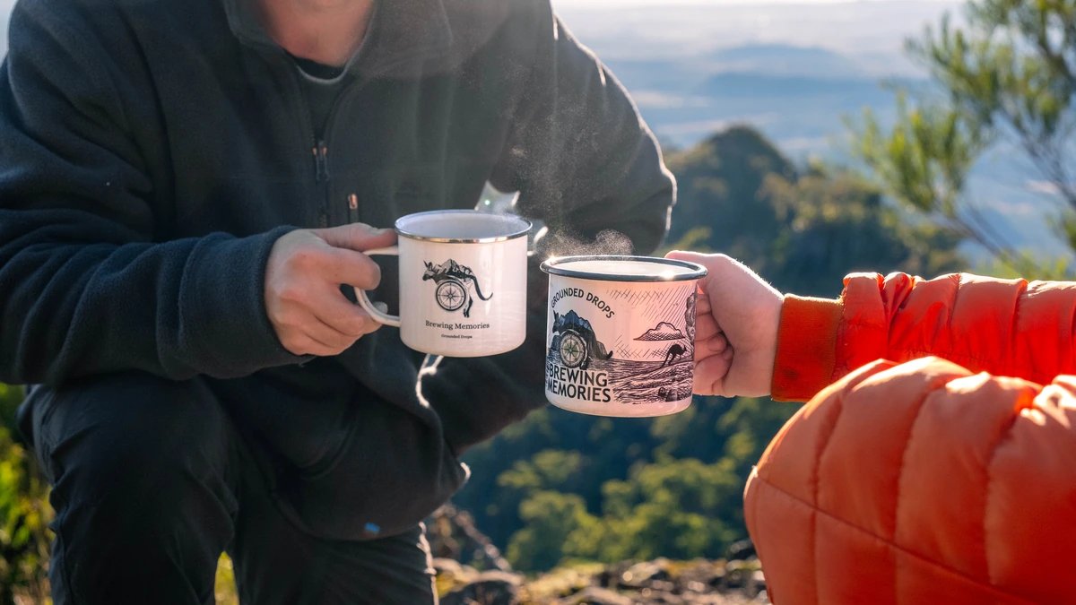Two people holding steaming Grounded Drops enamel coffee mugs at a bush lookout.