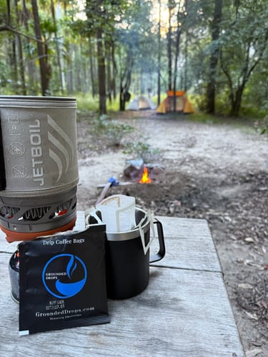 Drip coffee bag in a camp mug beside a camp stove with a campfire behind